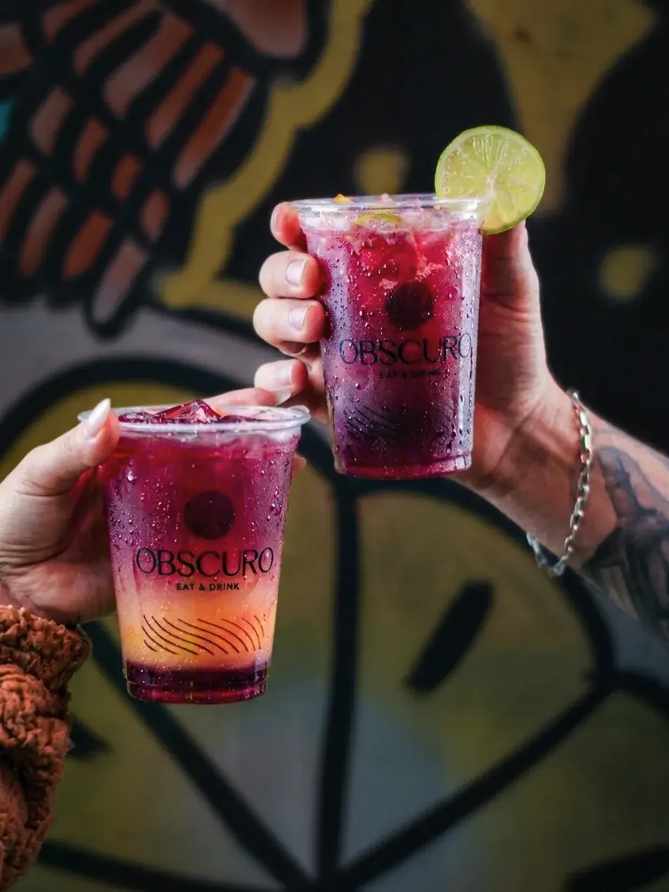 Duas mãos brindando com drinks roxos em copos do Obscuro